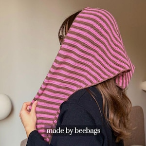 May include: A pink and brown striped crochet hood. The hood is made of a soft, textured yarn and features horizontal stripes. The text "made by beebags" is visible at the bottom of the image.