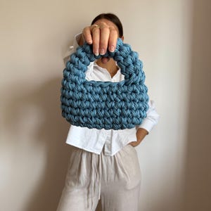 Może przedstawiać: Niebieska szydełkowa torebka z zaokrąglonym uchwytem. Torba wykonana jest z grubej, teksturowanej przędzy i ma strukturalny kształt. Torba jest trzymana przed osobą ubraną w białą koszulę i beżowe spodnie.