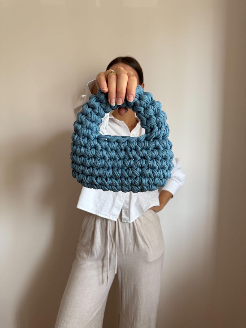 Może przedstawiać: Niebieska szydełkowa torebka z zaokrąglonym uchwytem. Torba wykonana jest z grubej, teksturowanej przędzy i ma strukturalny kształt. Torba jest trzymana przed osobą ubraną w białą koszulę i beżowe spodnie.