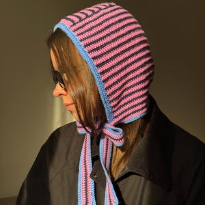 May include: A crochet bonnet featuring horizontal stripes in alternating shades of dark navy blue and light pink. The edges of the bonnet and the two long chin ties are finished with a solid light blue border, creating a distinct contrast.