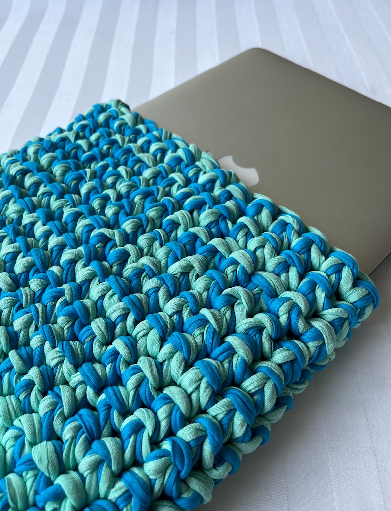 May include: A blue and green crocheted laptop sleeve. The sleeve is made of thick yarn and has a textured surface.