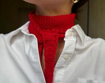 May include: A bright red, crocheted neck scarf is worn over a white button-down shirt. The scarf is tied in a knot, with the ends hanging down. Gold-colored oval earrings are visible.