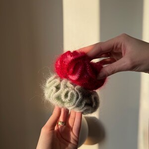 May include: Two handmade scrunchies, one red and one green, are held in front of a neutral background. The scrunchies appear to be made of a soft, fuzzy material. The red scrunchie is on top, and the green scrunchie is below it.