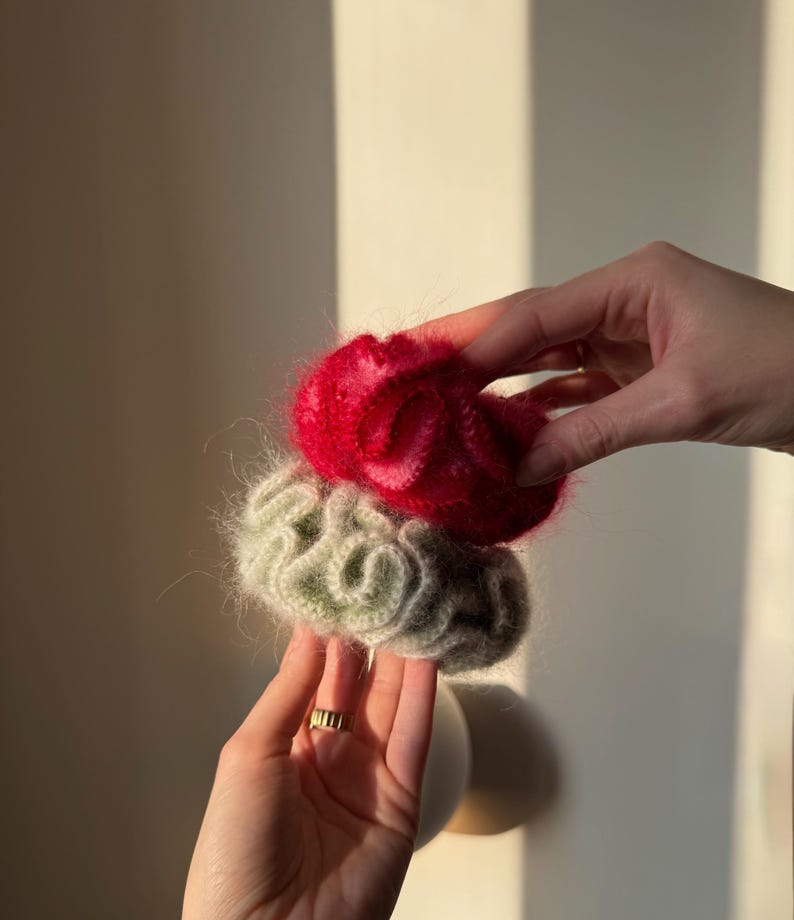 May include: Two handmade scrunchies, one red and one green, are held in front of a neutral background. The scrunchies appear to be made of a soft, fuzzy material. The red scrunchie is on top, and the green scrunchie is below it.