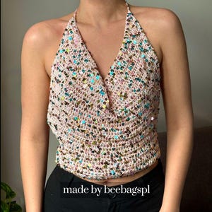 以下が含まれることがあります： ライトピンクのかぎ針編み素材で作られた、Vネックデザインのホルタートップ。カラフルなスパンコールで装飾されています。「made by beebagspl」の文字が下部に表示されています。
