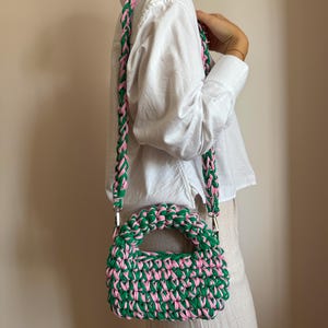 Może przedstawiać: Zielona i r&oacute;żowa szydełkowa torebka z pasującym paskiem. Torebka ma zaokrąglony kształt z uchwytem i tkaną fakturą. Pasek jest przymocowany za pomocą srebrnego osprzętu. Torebka jest noszona na ramieniu.