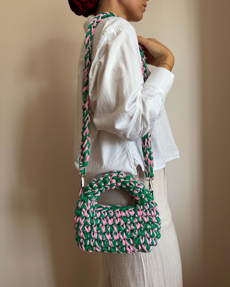 Może przedstawiać: Zielona i r&oacute;żowa szydełkowa torebka z pasującym paskiem. Torebka ma zaokrąglony kształt z uchwytem i tkaną fakturą. Pasek jest przymocowany za pomocą srebrnego osprzętu. Torebka jest noszona na ramieniu.