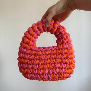 Może przedstawiać: Mała, ręcznie robiona torba typu tote w kolorze r&oacute;żowym i pomarańczowym. Torba wykonana jest z grubej, tkanej tkaniny, z zaokrąglonym uchwytem i strukturalnym kształtem. Połączenie kolor&oacute;w tworzy odważny, przyciągający wzrok dodatek. Wymiary: ok. 20 cm szerokości.