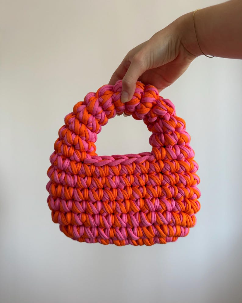 Może przedstawiać: Mała, ręcznie robiona torba typu tote w kolorze r&oacute;żowym i pomarańczowym. Torba wykonana jest z grubej, tkanej tkaniny, z zaokrąglonym uchwytem i strukturalnym kształtem. Połączenie kolor&oacute;w tworzy odważny, przyciągający wzrok dodatek. Wymiary: ok. 20 cm szerokości.