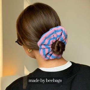 Könnte beinhalten: Ein blau-rosa gestreifter Häkel-Haargummi, der in einem Dutt getragen wird. Der Haargummi ist aus weichem Garn gefertigt und hat ein strukturiertes Aussehen. Der Text "made by beebags" ist unten im Bild zu sehen.