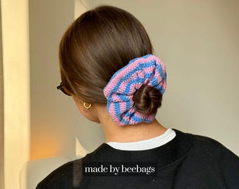 May include: A blue and pink striped crochet scrunchie worn in a bun. The scrunchie is made of soft yarn and has a textured appearance. The text "made by beebags" is visible at the bottom of the image.