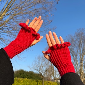 May include: Bright red fingerless gloves with a ruffled edge. The gloves are knitted and appear to be made of a soft yarn. The background shows a clear blue sky and bare tree branches.