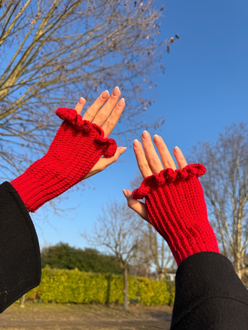 May include: Bright red fingerless gloves with a ruffled edge. The gloves are knitted and appear to be made of a soft yarn. The background shows a clear blue sky and bare tree branches.