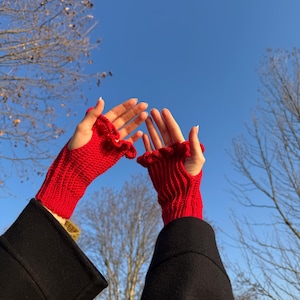 May include: Bright red fingerless gloves with a ruffled edge are held up against a clear blue sky. The gloves are made of a knitted material and are worn with a black coat. The gloves are a vibrant accessory.