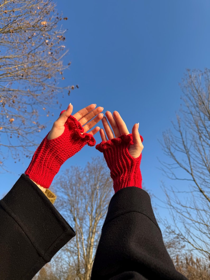 May include: Bright red fingerless gloves with a ruffled edge are held up against a clear blue sky. The gloves are made of a knitted material and are worn with a black coat. The gloves are a vibrant accessory.