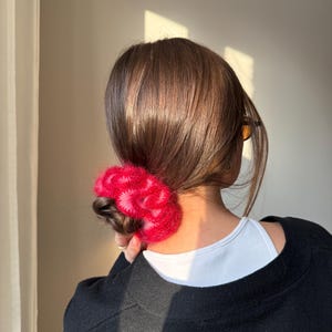 May include: A person's hair is styled in a low bun, secured with a vibrant, fluffy, red scrunchie. The scrunchie adds a pop of color against the dark hair and black sweater. The person is wearing a white top underneath the sweater.