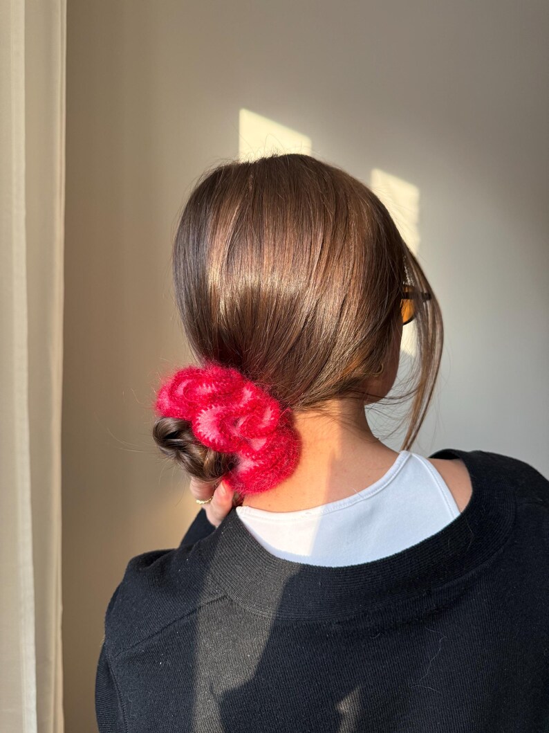 May include: A person's hair is styled in a low bun, secured with a vibrant, fluffy, red scrunchie. The scrunchie adds a pop of color against the dark hair and black sweater. The person is wearing a white top underneath the sweater.