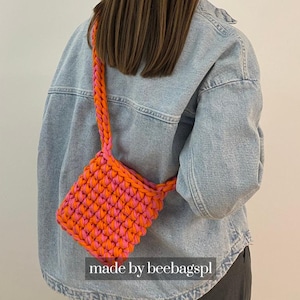 May include: A small, handmade, crocheted crossbody bag in vibrant orange and pink. The bag features a braided strap and a textured stitch pattern. The text "made by beebagspl" is visible at the bottom of the image.