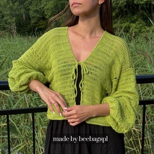 May include: A lime green crochet cardigan featuring long, voluminous sleeves, a deep V-neckline, and a front tie closure. The garment has a textured, open-stitch pattern and is cropped at the waist. The text made by beebagspl is visible.
