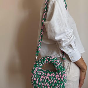 Może przedstawiać: Zielono-r&oacute;żowa tkana torebka z pasującym plecionym paskiem. Torebka ma zaokrąglony uchwyt i prostokątny kształt. Pasek jest przymocowany za pomocą srebrnego osprzętu. Torebka jest noszona na ramieniu.