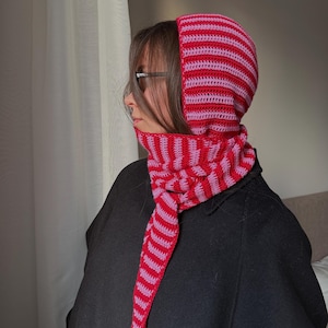 Puede incluir: Un conjunto de capucha y bufanda de crochet a rayas rojas y rosas. La capucha y la bufanda est&aacute;n hechas de un hilo suave y texturizado. La capucha cubre la cabeza y el cuello, con una bufanda que cuelga por delante.