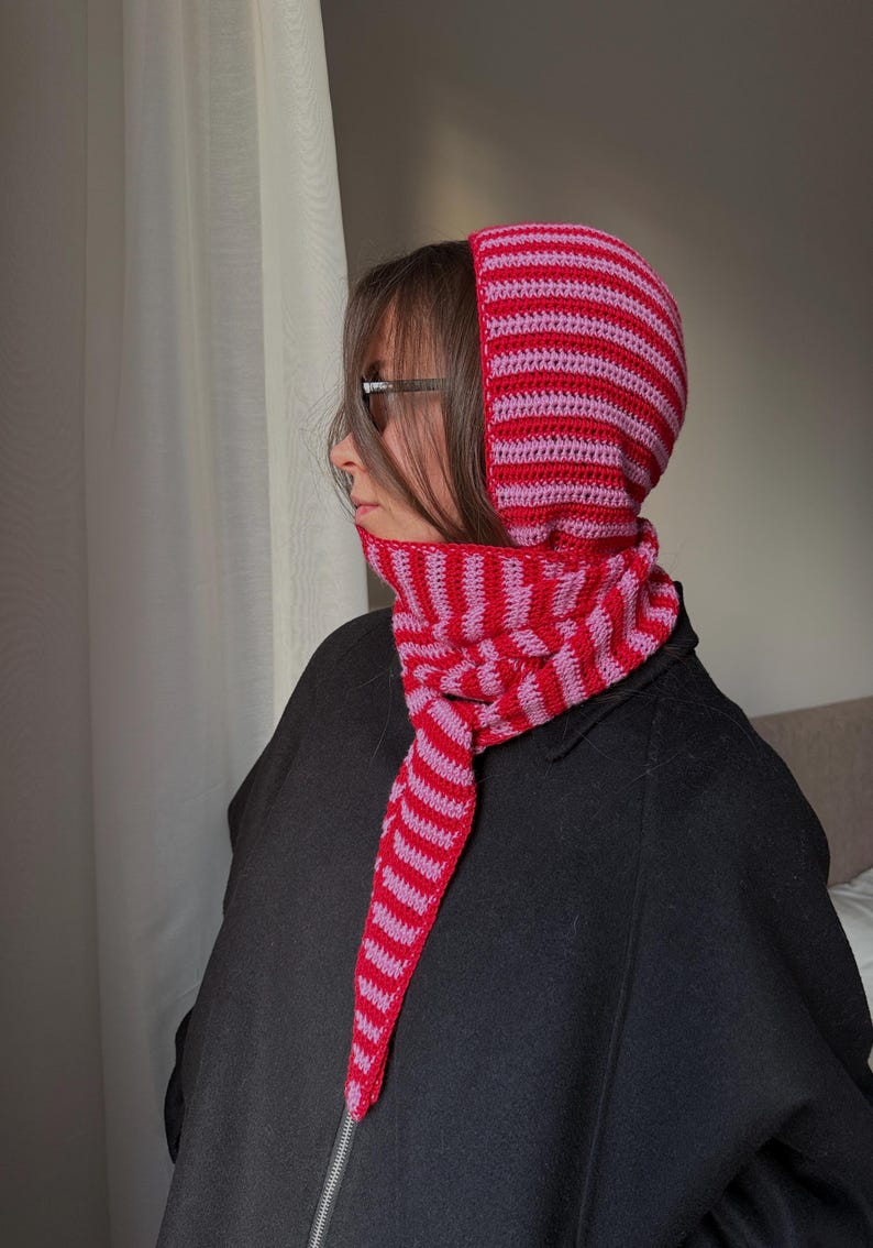 Puede incluir: Un conjunto de capucha y bufanda de crochet a rayas rojas y rosas. La capucha y la bufanda est&aacute;n hechas de un hilo suave y texturizado. La capucha cubre la cabeza y el cuello, con una bufanda que cuelga por delante.