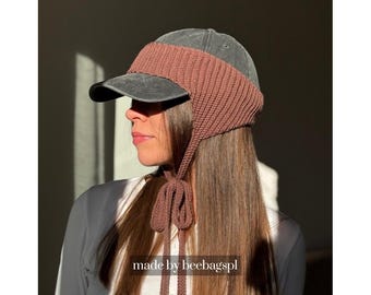 May include: A charcoal gray baseball cap featuring a wide, ribbed, brown knitted ear warmer band that wraps around the crown and secures under the chin with two long, matching knitted ties. The text made by beebagspl is visible.