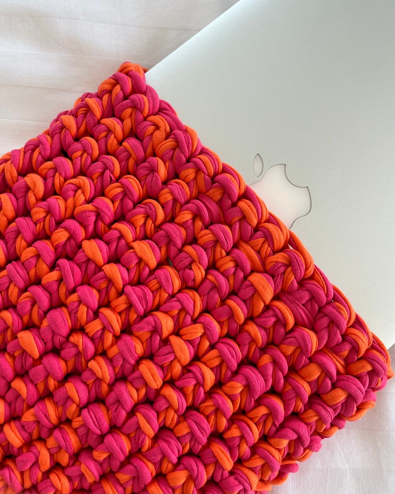 May include: A crocheted laptop sleeve in pink and orange. The sleeve is made with thick yarn and has a simple, textured design.