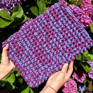 May include: A crocheted bag made with two shades of purple yarn. The bag is held in a person's hands against a background of purple flowers.
