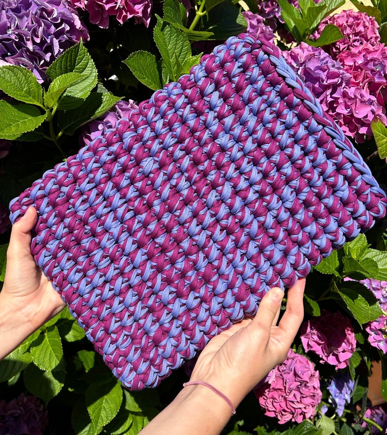 May include: A crocheted bag made with two shades of purple yarn. The bag is held in a person's hands against a background of purple flowers.