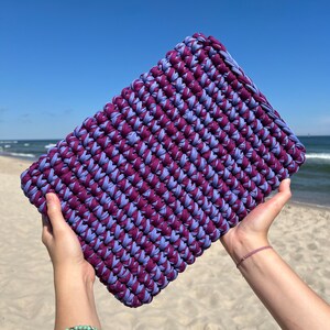 May include: A purple and blue crocheted clutch bag held in the air against a blue sky and sandy beach background.
