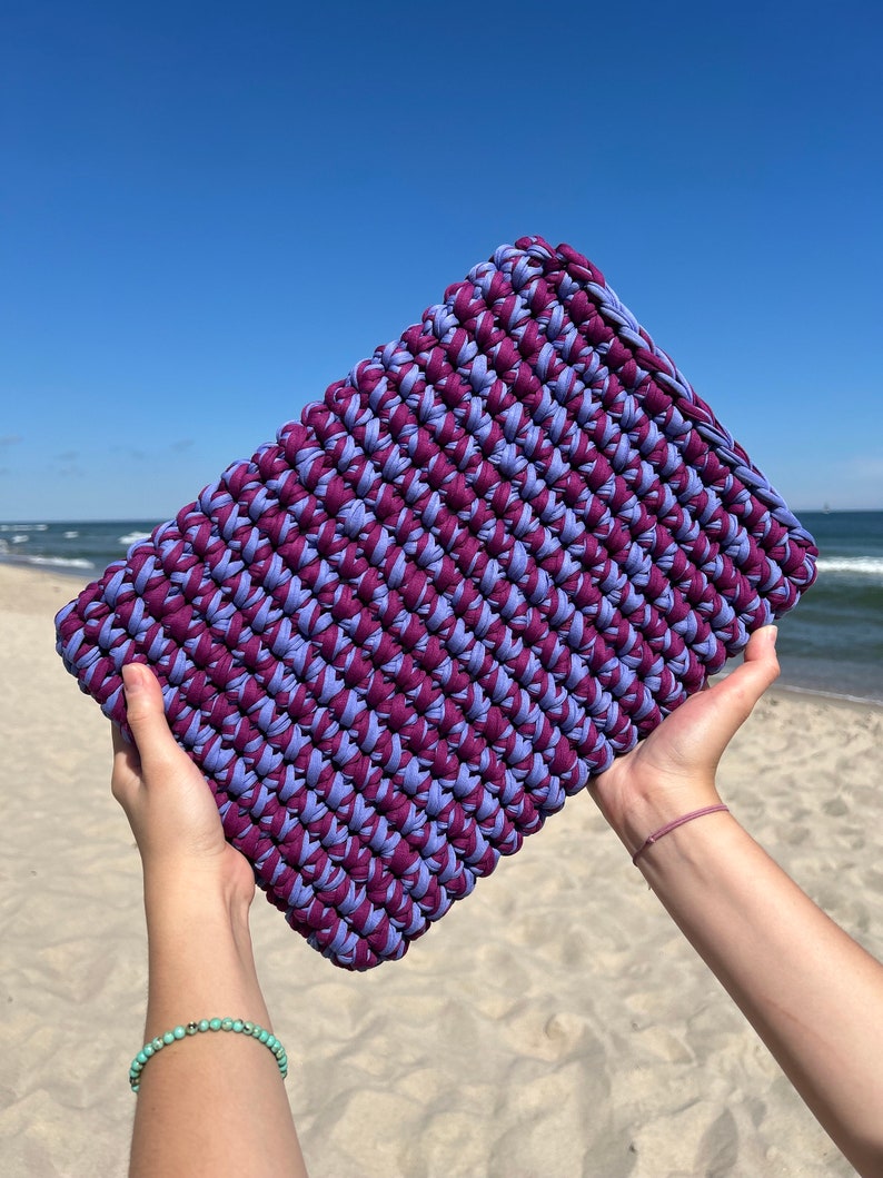 May include: A purple and blue crocheted clutch bag held in the air against a blue sky and sandy beach background.