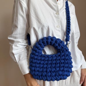 Może przedstawiać: Ręcznie robiona, kobaltowa torba z szydełka z plecionym paskiem na ramię. Torba ma zaokrąglony kształt z małym uchwytem i srebrnymi elementami. Torba jest wykonana z grubej, teksturowanej przędzy.