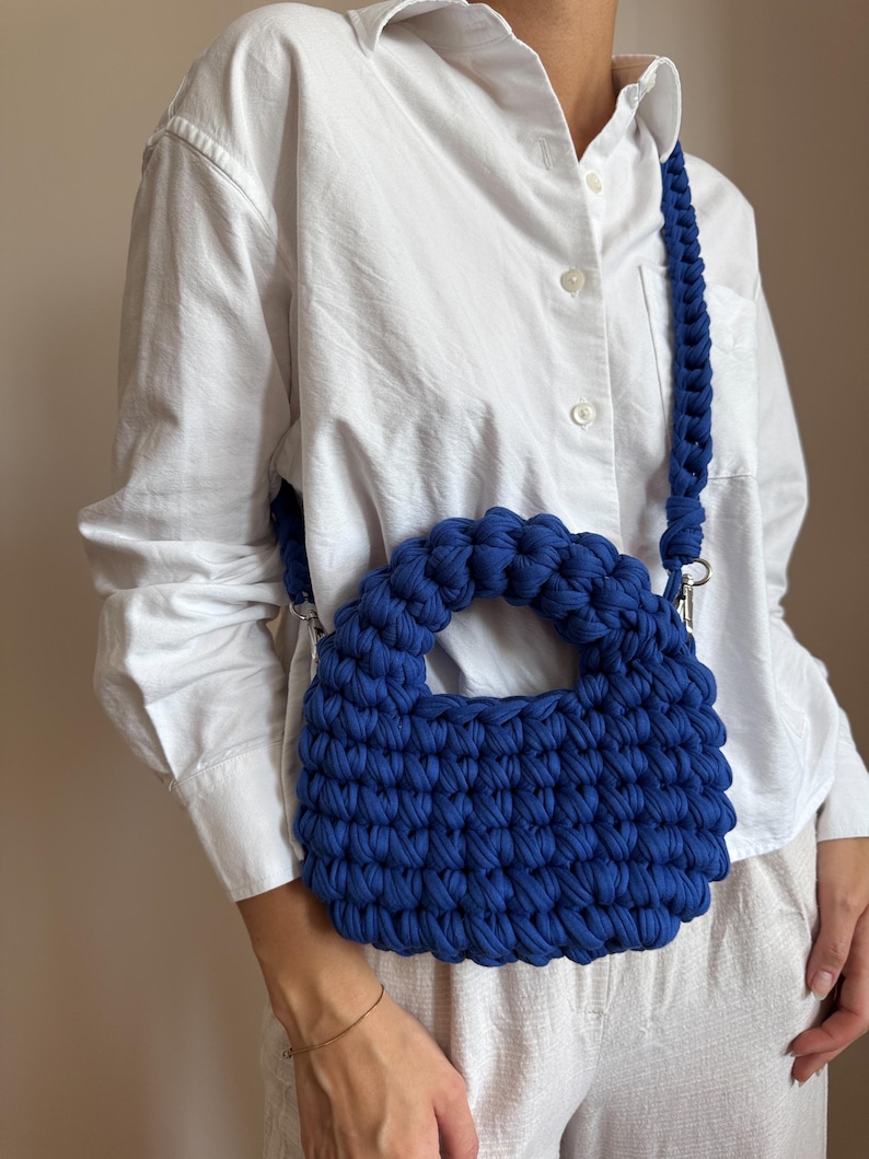 Może przedstawiać: Ręcznie robiona, kobaltowa torba z szydełka z plecionym paskiem na ramię. Torba ma zaokrąglony kształt z małym uchwytem i srebrnymi elementami. Torba jest wykonana z grubej, teksturowanej przędzy.