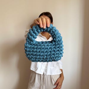 Może przedstawiać: Niebieska szydełkowa torebka z zaokrąglonym uchwytem. Torba wykonana jest z grubej, teksturowanej przędzy i ma kompaktowy, strukturalny kształt. Kolor jest stonowany, zakurzony niebieski, a torba wydaje się być ręcznie robiona.