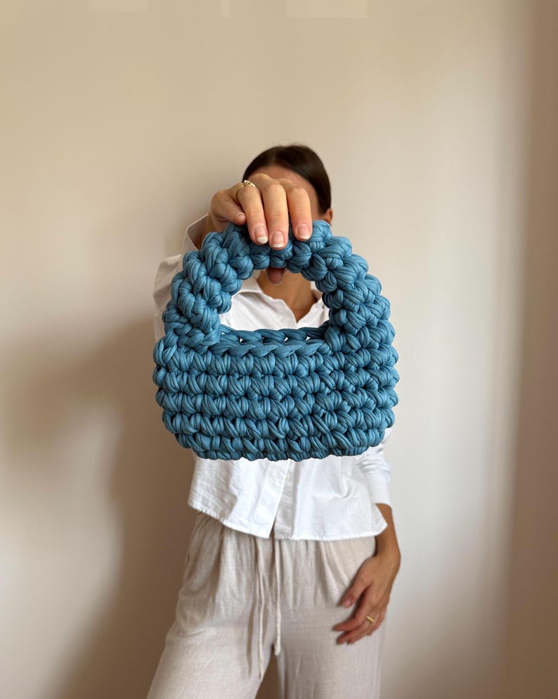 Może przedstawiać: Niebieska szydełkowa torebka z zaokrąglonym uchwytem. Torba wykonana jest z grubej, teksturowanej przędzy i ma kompaktowy, strukturalny kształt. Kolor jest stonowany, zakurzony niebieski, a torba wydaje się być ręcznie robiona.