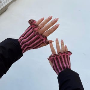 May include: Crocheted fingerless gloves in pink and brown stripes with a ruffled edge. The gloves are worn against a white background, with the wearer's black jacket sleeves visible.