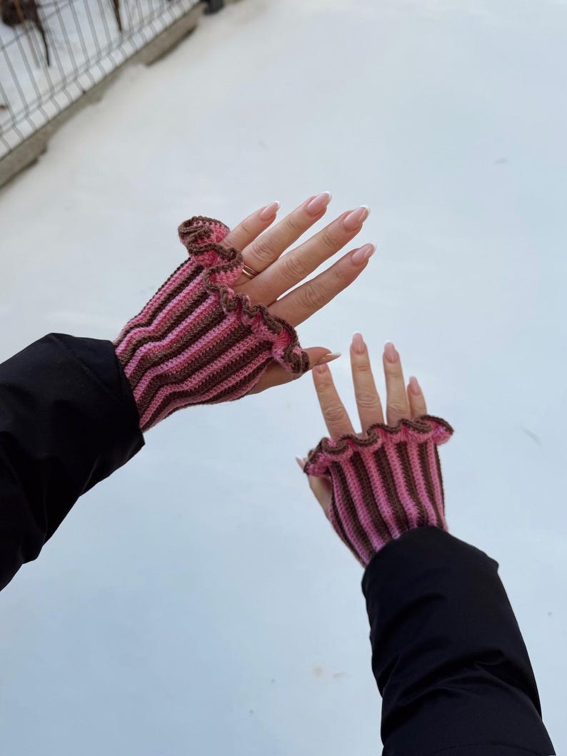 May include: Crocheted fingerless gloves in pink and brown stripes with a ruffled edge. The gloves are worn against a white background, with the wearer's black jacket sleeves visible.