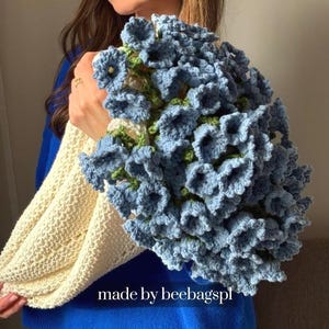 Op de afbeelding: Een handgemaakt gehaakt boeket van blauwe bloemen met groene accenten, gehouden in een crèmekleurige gehaakte wikkel. Het boeket is groot en gedetailleerd, met een textuur. De tekst "made by beebagspl" is zichtbaar.