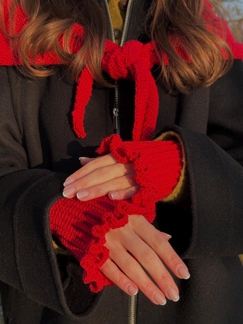 May include: A black coat with a red crocheted scarf and matching wrist warmers. The wrist warmers have a ruffled edge. The scarf is tied in a bow. The coat has a zipper closure.