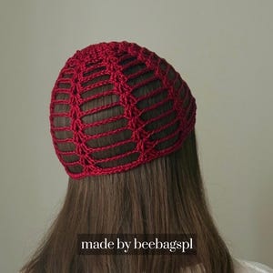 Peut inclure: Un bonnet rouge en crochet avec un motif ajouré en forme de toile d'araignée. Le bonnet est porté sur la tête d'une personne, avec de longs cheveux bruns visibles en dessous. Le texte "made by beebagspl" est en bas de l'image.