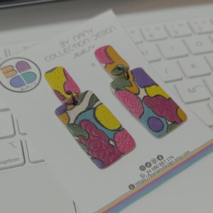 May include: A pair of colorful dangle earrings with a geometric pattern. The earrings are made of polymer clay and have a white background with pink, yellow, blue, and purple accents. The earrings are hanging from a white card with the text "BY NANY COLLECTION DESIGN JEWELRY" and contact information.