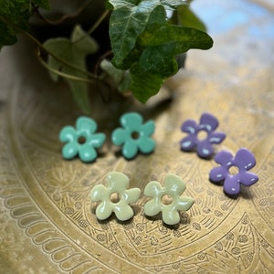 May include: A collection of flower-shaped stud earrings in various pastel colors: mint green, pale yellow, and lavender. The earrings are arranged on a decorative, etched brass surface, with green foliage in the background.