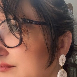 May include: A pair of white and brown patterned dangle earrings with a gold chain. The earrings are made of two round discs, one larger than the other.
