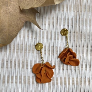 May include: A pair of dangle earrings with a gold chain and a cluster of orange flower-shaped charms. The earrings are on a woven background.