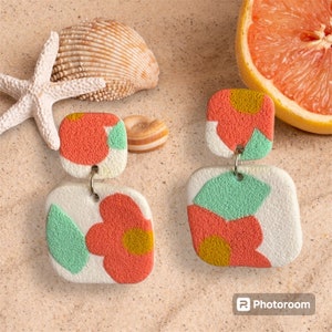 May include: A pair of square, dangle earrings with a textured, abstract design. The earrings feature a white base with coral, yellow, and mint green shapes. The earrings are displayed on a sandy surface with seashells and a grapefruit.