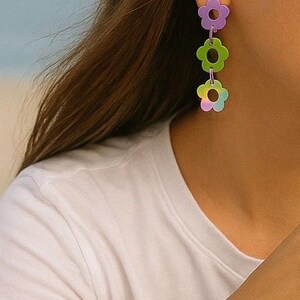 May include: A woman wearing a white t-shirt and a green, white, and orange crocheted hat. She is wearing a flower-shaped bracelet and earrings with purple, green, and orange flowers.