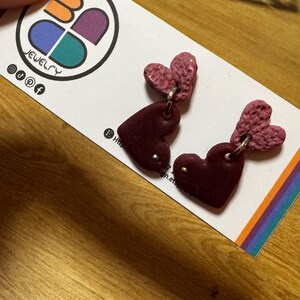 May include: A pair of heart-shaped earrings. The earrings feature a dark red heart pendant with a textured, light pink heart above. The earrings are displayed on a white card with a colorful logo and the word "JEWELRY".