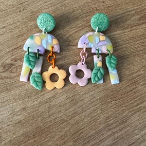 May include: A pair of colorful dangle earrings with a geometric design. The earrings feature a green, pink, and yellow color scheme with a floral motif.