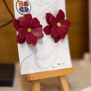 May include: A pair of burgundy flower earrings with gold centers displayed on a small wooden easel. The earrings are on a white card with a logo and a line drawing of a woman's neck and necklace. The earrings are part of the BA Jewelry Collection.